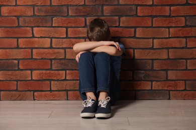 Child abuse. Upset boy sitting on floor near brick wall indoors