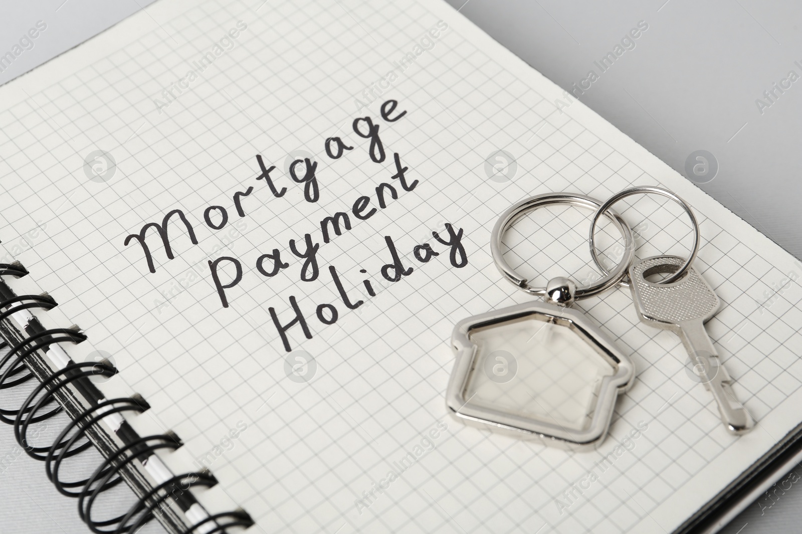 Photo of Key with trinket in shape of house and phrase Mortgage payment holiday written on notebook against light grey background, closeup