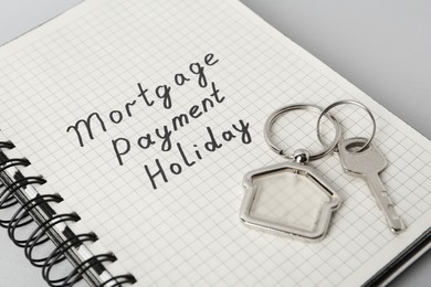 Photo of Key with trinket in shape of house and phrase Mortgage payment holiday written on notebook against light grey background, closeup