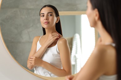 Photo of Suffering from allergy. Young woman scratching her skin near mirror in bathroom