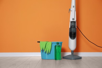 Photo of Modern steam mop, bucket with gloves and bottle of cleaning product on floor near orange wall, space for text
