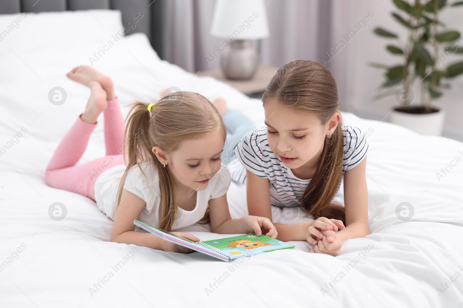 Photo of Cute little sisters reading book together on bed at home