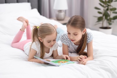 Cute little sisters reading book together on bed at home