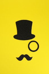 Photo of Man's face made of fake mustache, hat and monocle on yellow background, top view