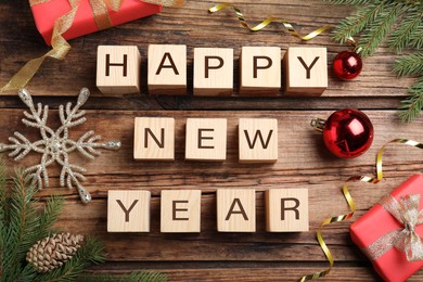 Photo of Happy New Year greeting card. Phrase made with cubes and festive decor on wooden background, flat lay