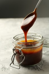 Taking tasty barbeque sauce from jar with spoon at grey textured table, closeup