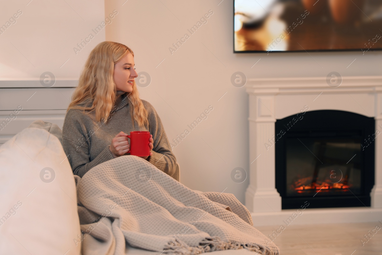 Photo of Young woman with cup of hot drink resting near fireplace at home