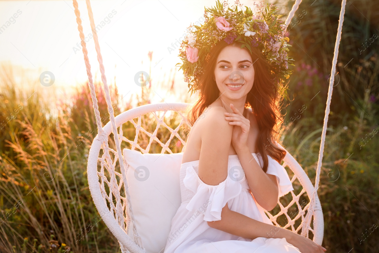 Photo of Young woman wearing wreath made of beautiful flowers on swing chair outdoors at sunset