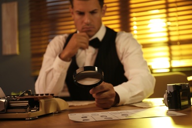 Photo of Old fashioned detective with magnifying glass working at table in office, focus on hand