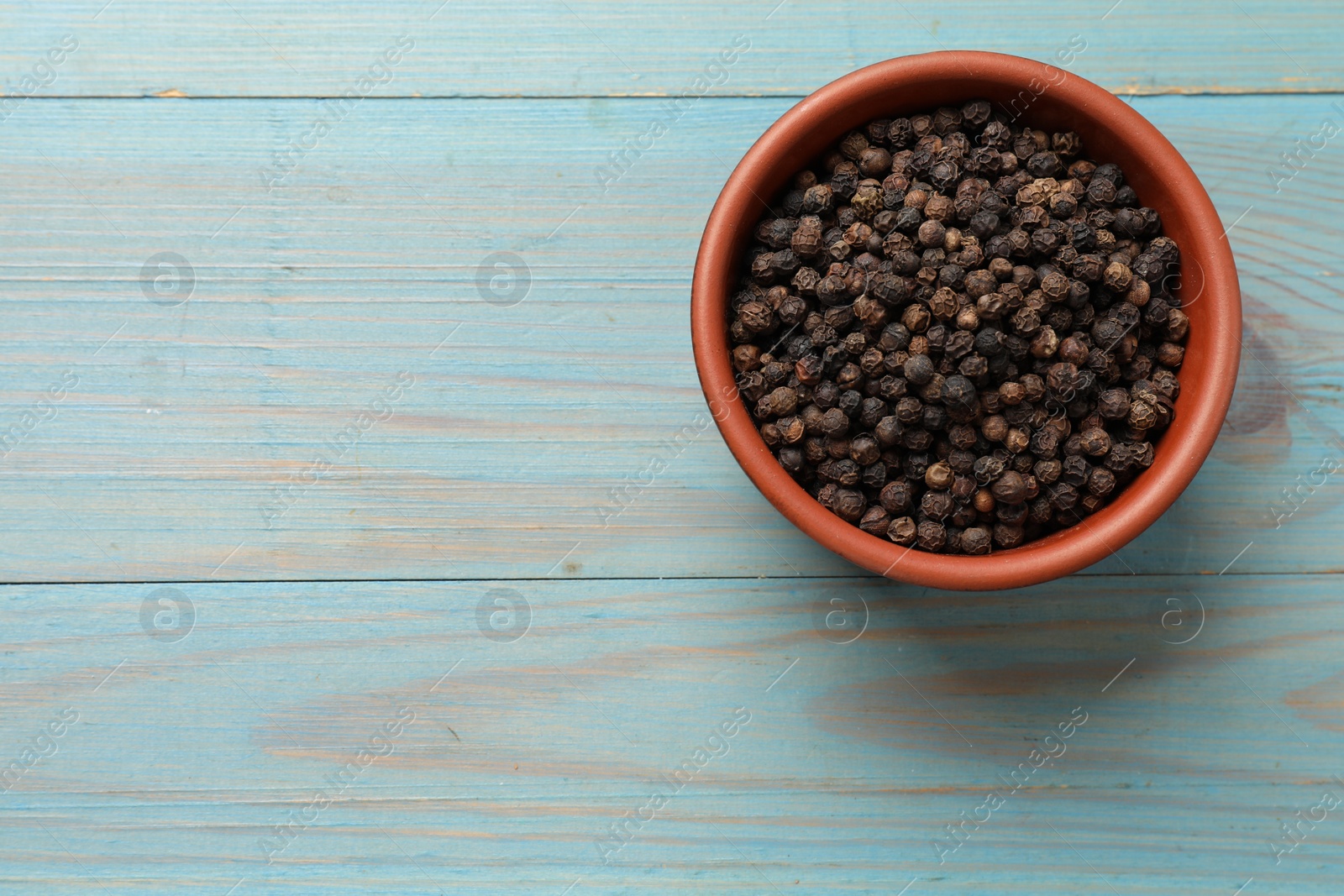 Photo of Aromatic spice. Black pepper in bowl on light blue wooden table, top view. Space for text