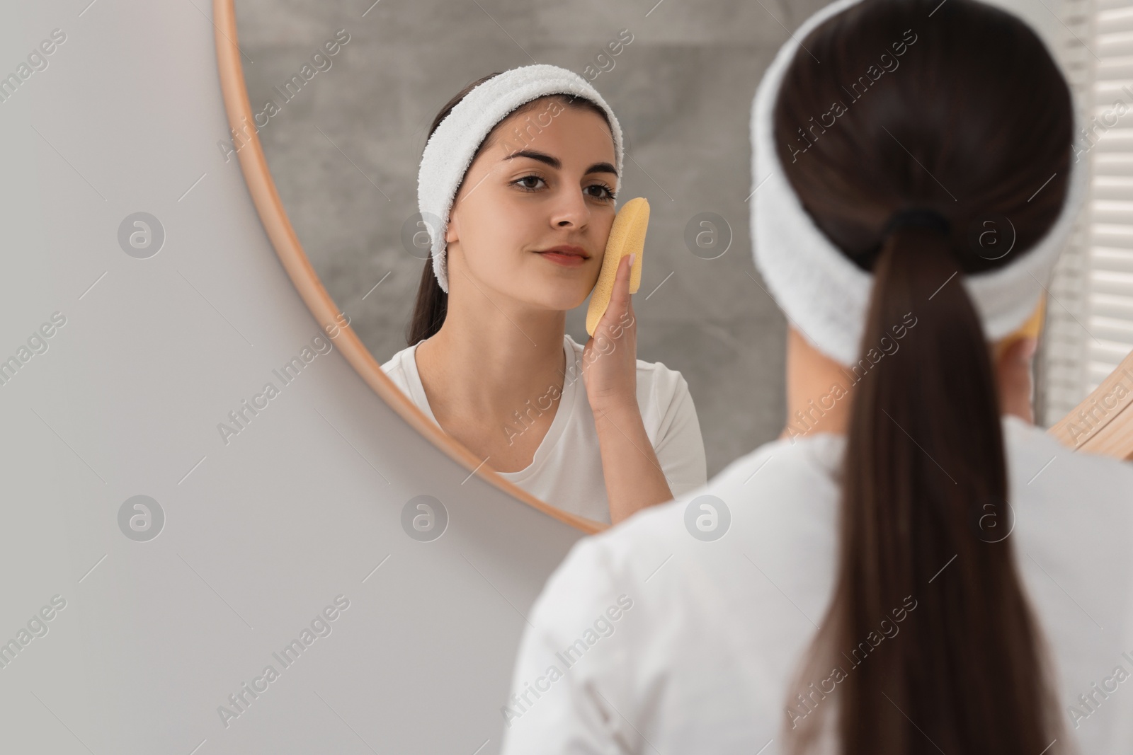 Photo of Young woman with headband washing her face using sponge near mirror in bathroom