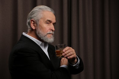 Photo of Senior man in suit holding glass of whiskey with ice cubes on brown background. Space for text