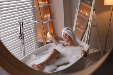 Photo of Beautiful woman taking bath in tub with foam indoors, reflection in mirror. Romantic atmosphere