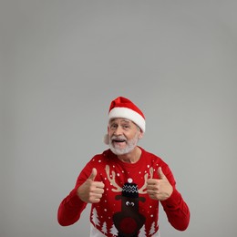 Photo of Senior man in Christmas sweater and Santa hat showing thumbs up on grey background