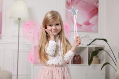 Photo of Cute little girl in fairy costume with pink wings and magic wand at home