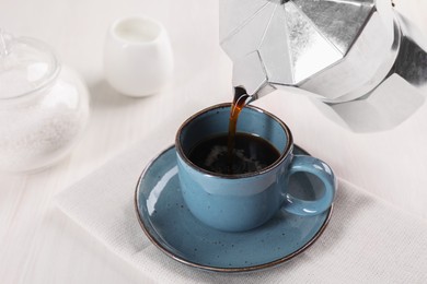 Pouring aromatic coffee from moka pot into cup at white wooden table, closeup