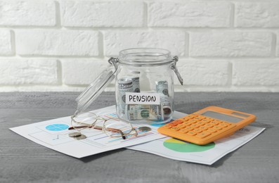 Photo of Glass jar with word Pension, money, calculator, charts and glasses on grey wooden table
