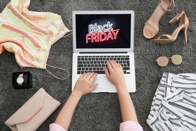 Photo of Woman surrounded by clothes and accessories using laptop with Black Friday announcement on grey carpet, top view