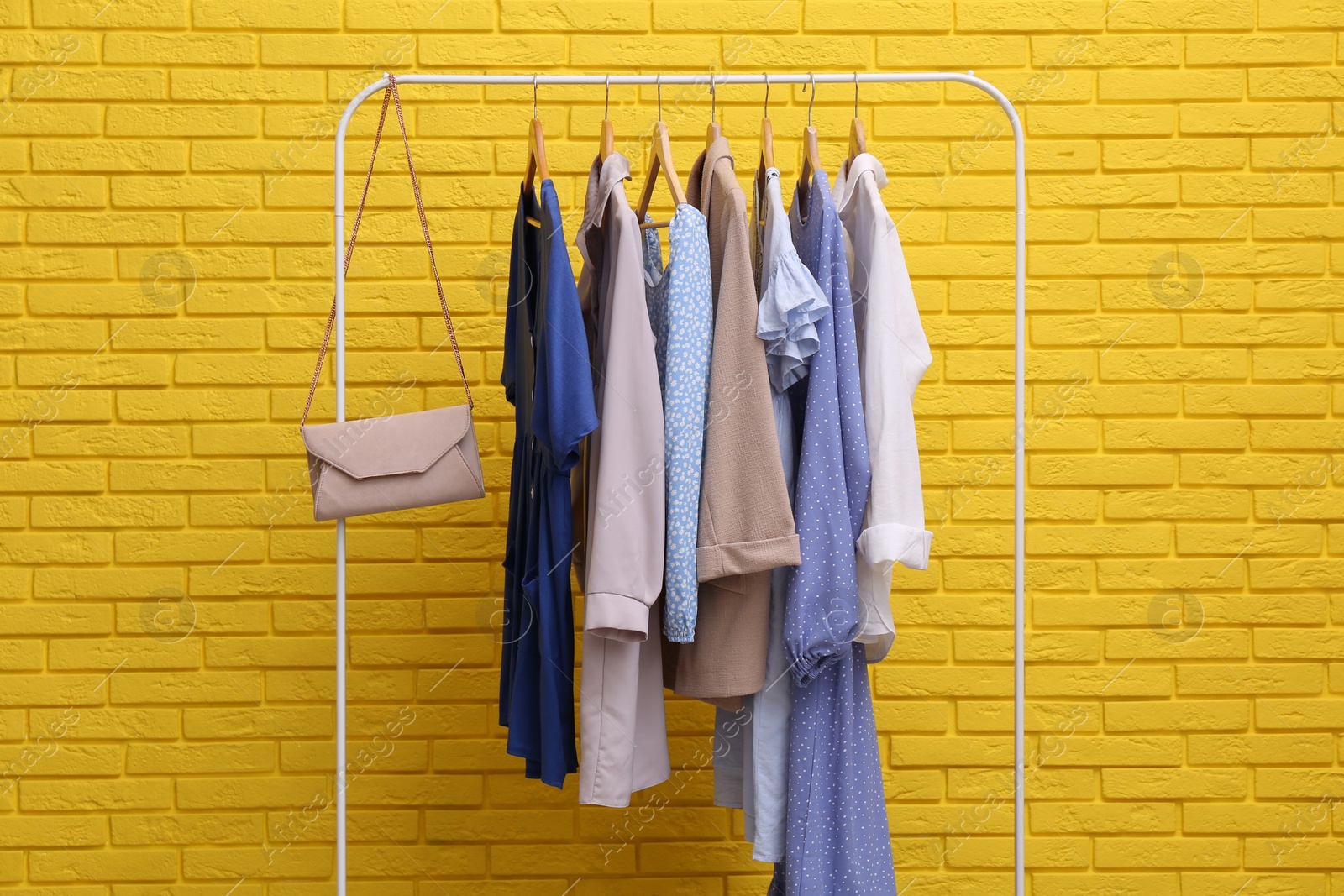 Photo of Rack with different stylish clothes and bag near yellow brick wall
