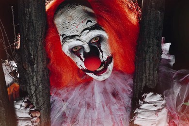 Photo of Terrifying clown hiding behind trees outdoors at night, closeup. Halloween party costume