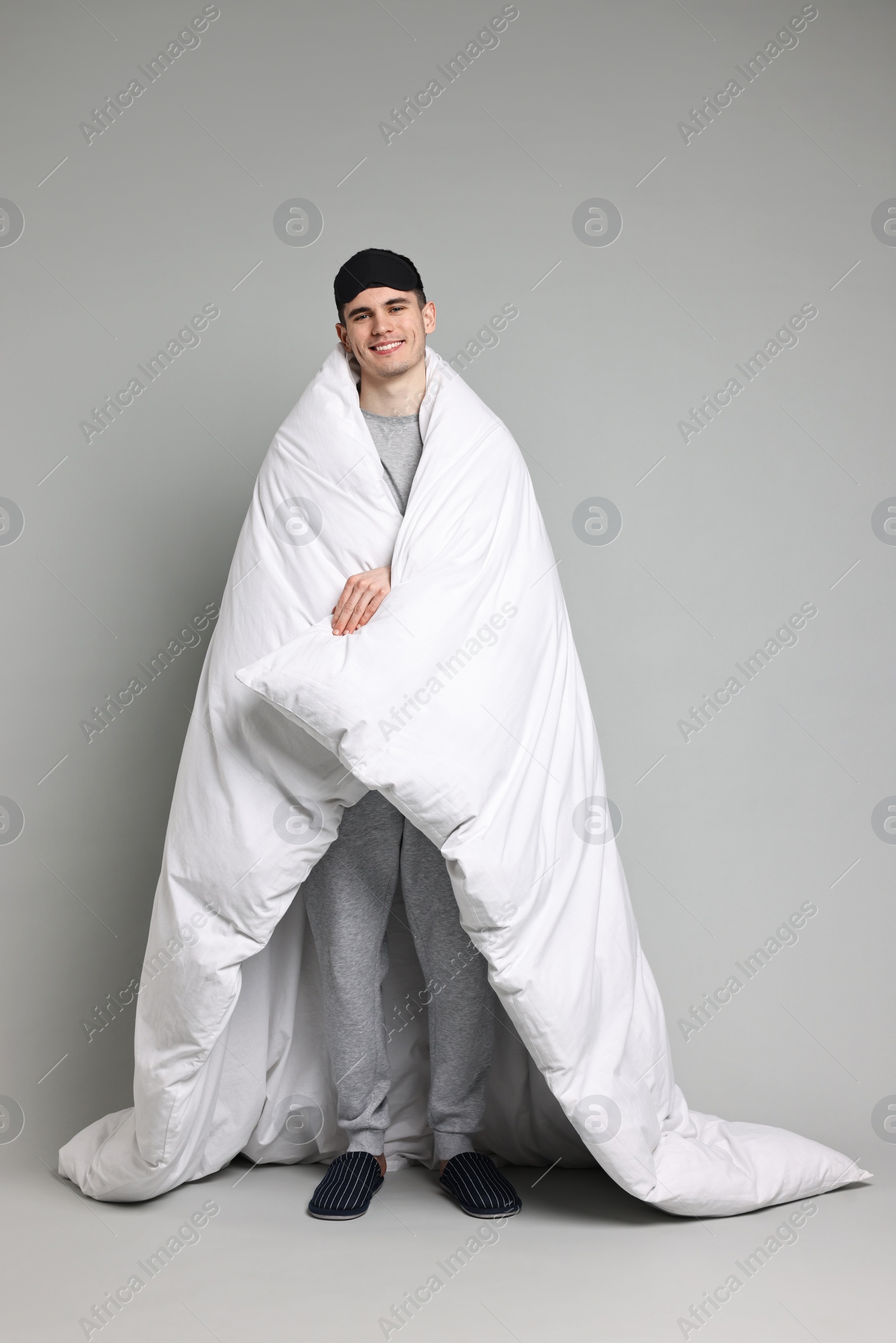 Photo of Man in pyjama and sleep mask wrapped in blanket on grey background