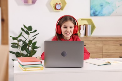 E-learning. Cute girl using laptop and headphones during online lesson at table indoors
