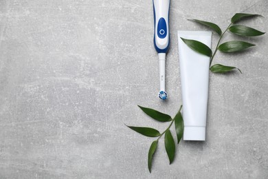 Photo of Electric toothbrush, tube of paste and leaves on grey textured table, flat lay. Space for text