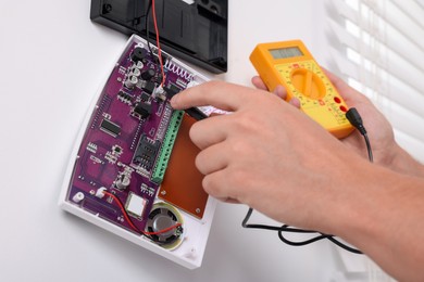 Photo of Man using digital multimeter while installing home security alarm system on white wall indoors, closeup