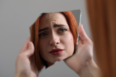 Photo of Young woman looking at herself in shard of broken mirror on light grey background, closeup