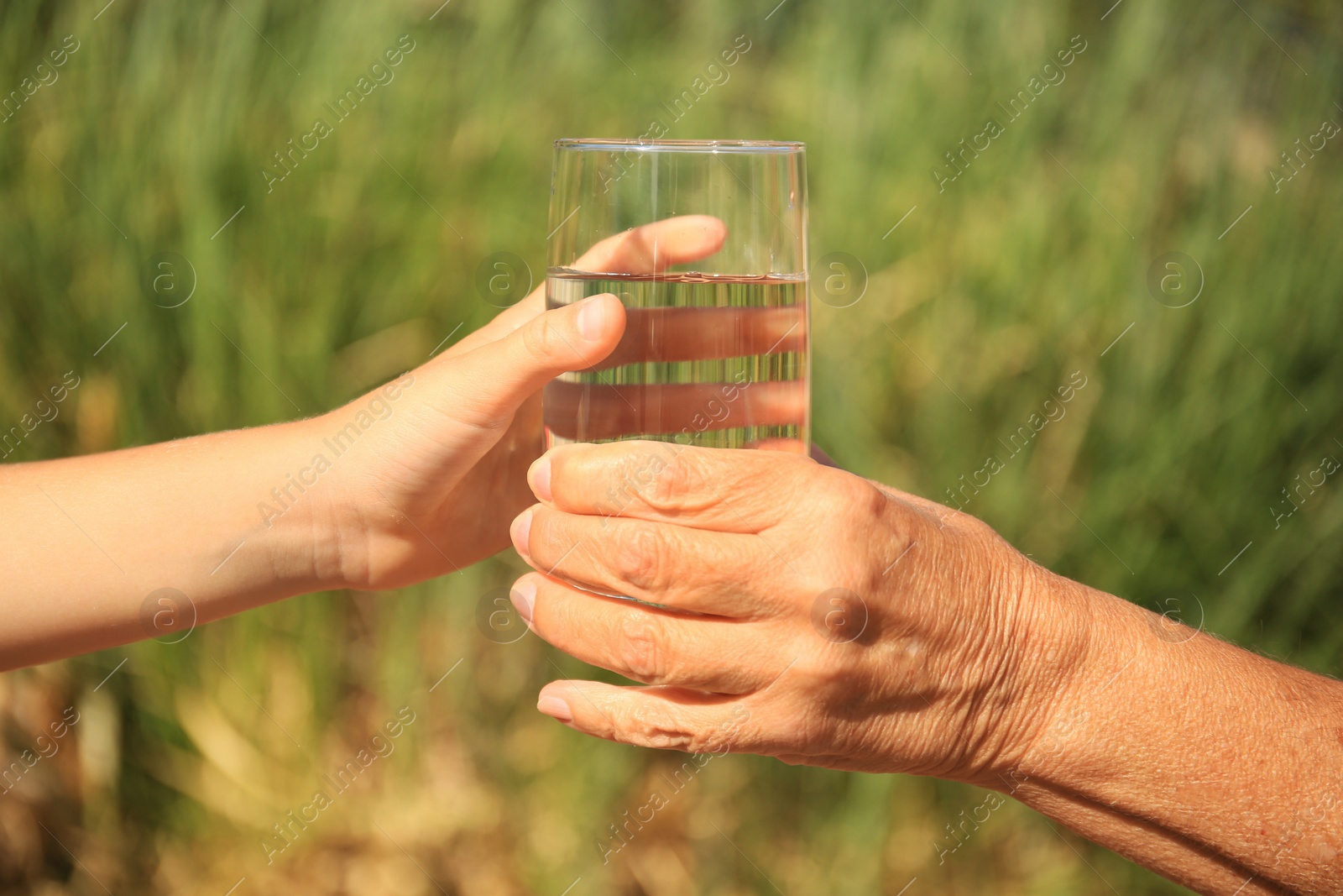 Photo of Child giving glass of water to elderly woman outdoors on sunny day, closeup