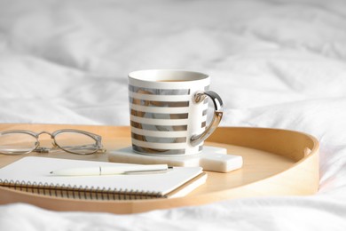 Photo of Mug of hot drink with stylish cup coaster, glasses and notebook on bed in room