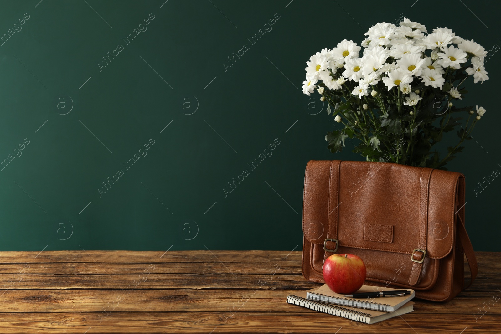 Photo of Leather briefcase, apple, stationery and flowers on wooden table near chalkboard, space for text. Teacher's Day