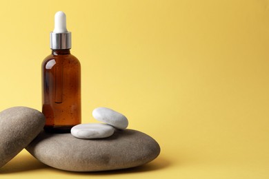 Photo of Bottle of face serum and spa stones on yellow background, space for text
