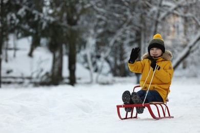 Photo of Cute little boy enjoying sledge ride through snow in winter park, space for text