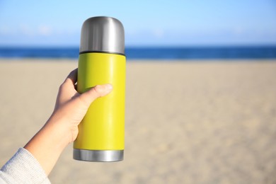 Woman holding yellow thermos with hot drink on beach near sea, closeup. Space for text