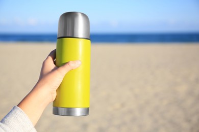 Photo of Woman holding yellow thermos with hot drink on beach near sea, closeup. Space for text