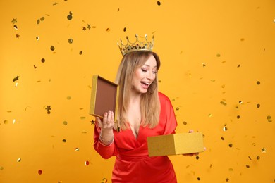 Happy young woman in party crown with gift box and confetti on yellow background