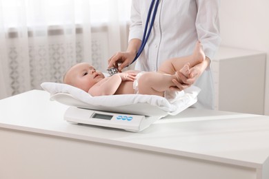 Photo of Pediatrician weighting and examining cute little baby with stethoscope in clinic, closeup