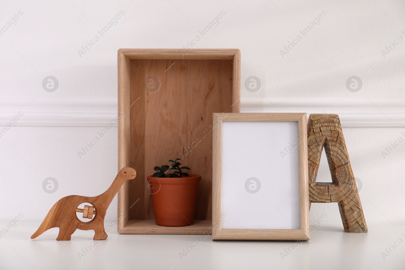 Photo of Empty photo frame, cute toy dinosaur, wooden letter and crate with houseplant near wall, space for text. Baby room interior element