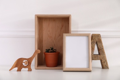 Photo of Empty photo frame, cute toy dinosaur, wooden letter and crate with houseplant near wall, space for text. Baby room interior element