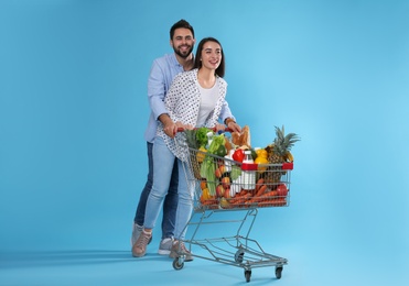 Photo of Young couple with shopping cart full of groceries on light blue background