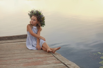 Cute little girl wearing wreath made of beautiful flowers on pier near river