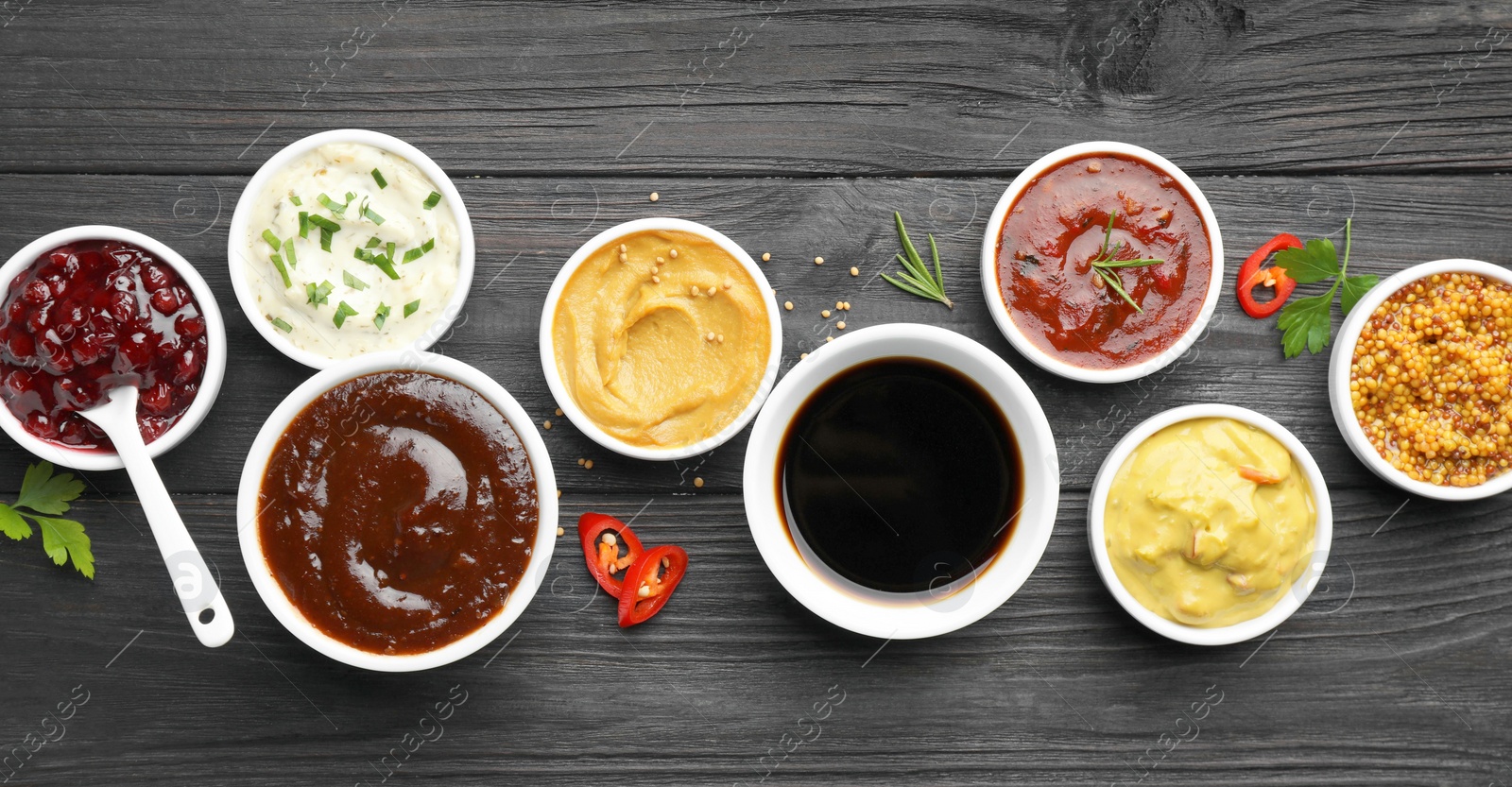 Photo of Different tasty sauces in bowls, parsley, chili pepper and rosemary on black wooden table, flat lay