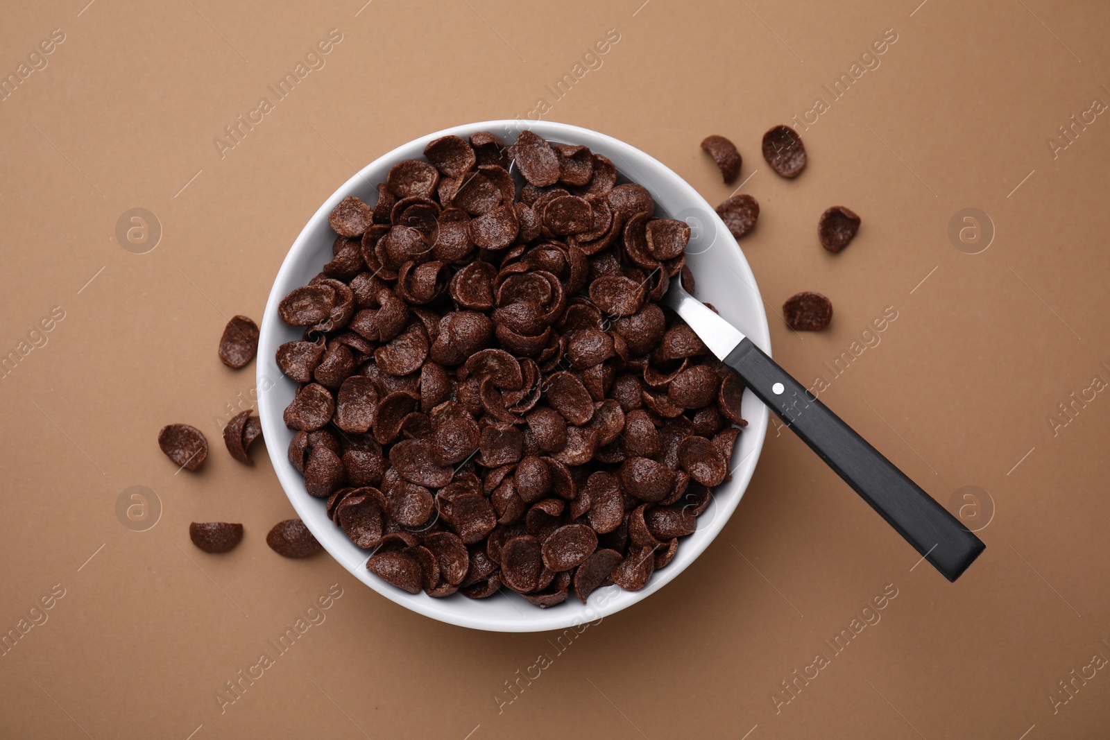 Photo of Breakfast cereal. Chocolate corn flakes in bowl and spoon on brown table, top view