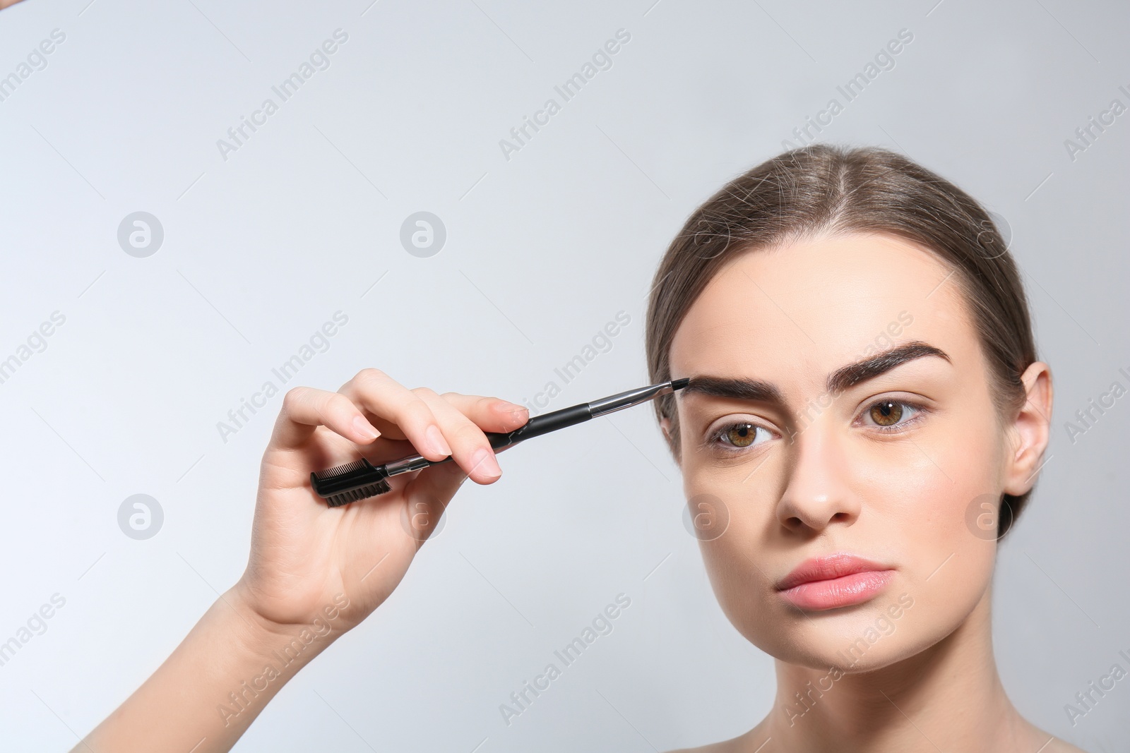 Photo of Young woman correcting shape of eyebrow with brush on light background