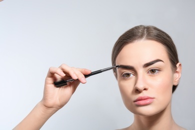 Photo of Young woman correcting shape of eyebrow with brush on light background