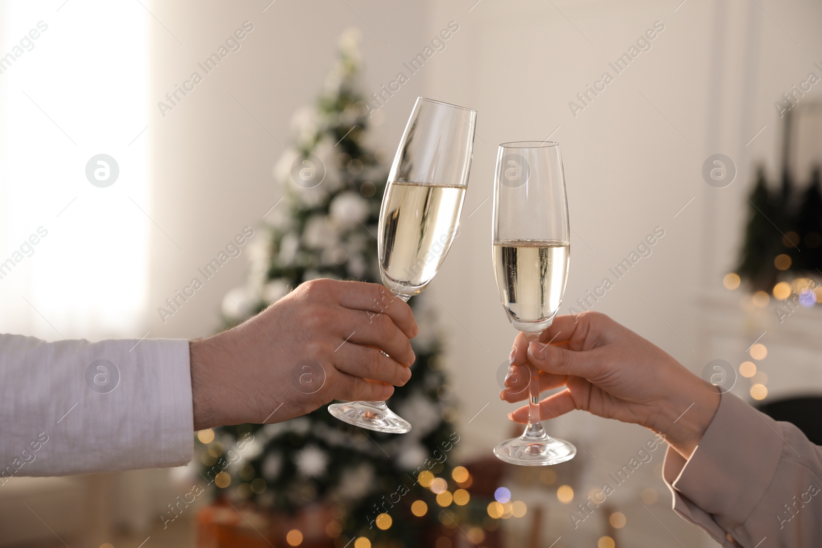 Photo of Couple clinking glasses of champagne in room decorated for Christmas, closeup. Holiday cheer and drink