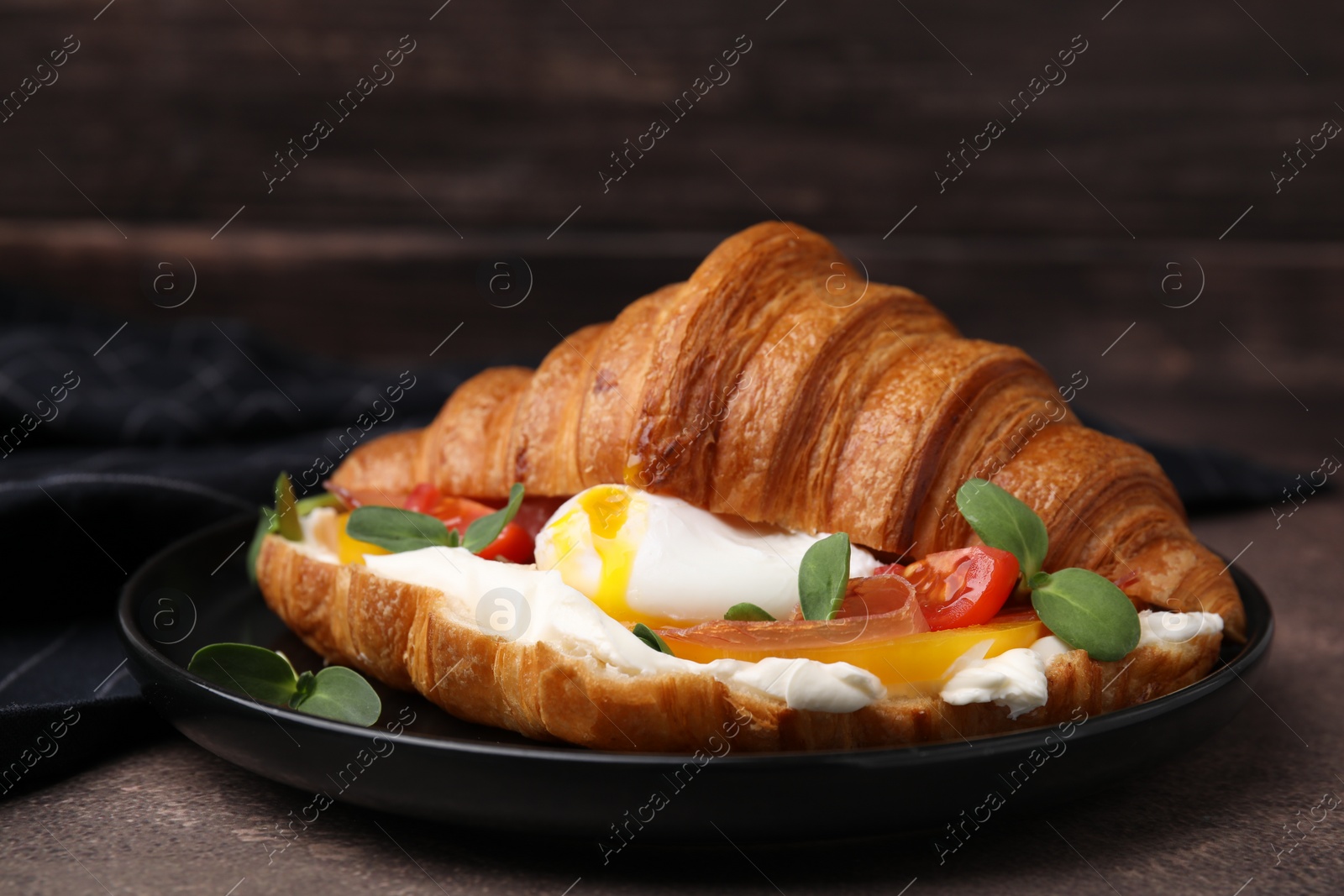 Photo of Tasty croissant with fried egg, tomato and microgreens on brown textured table, closeup