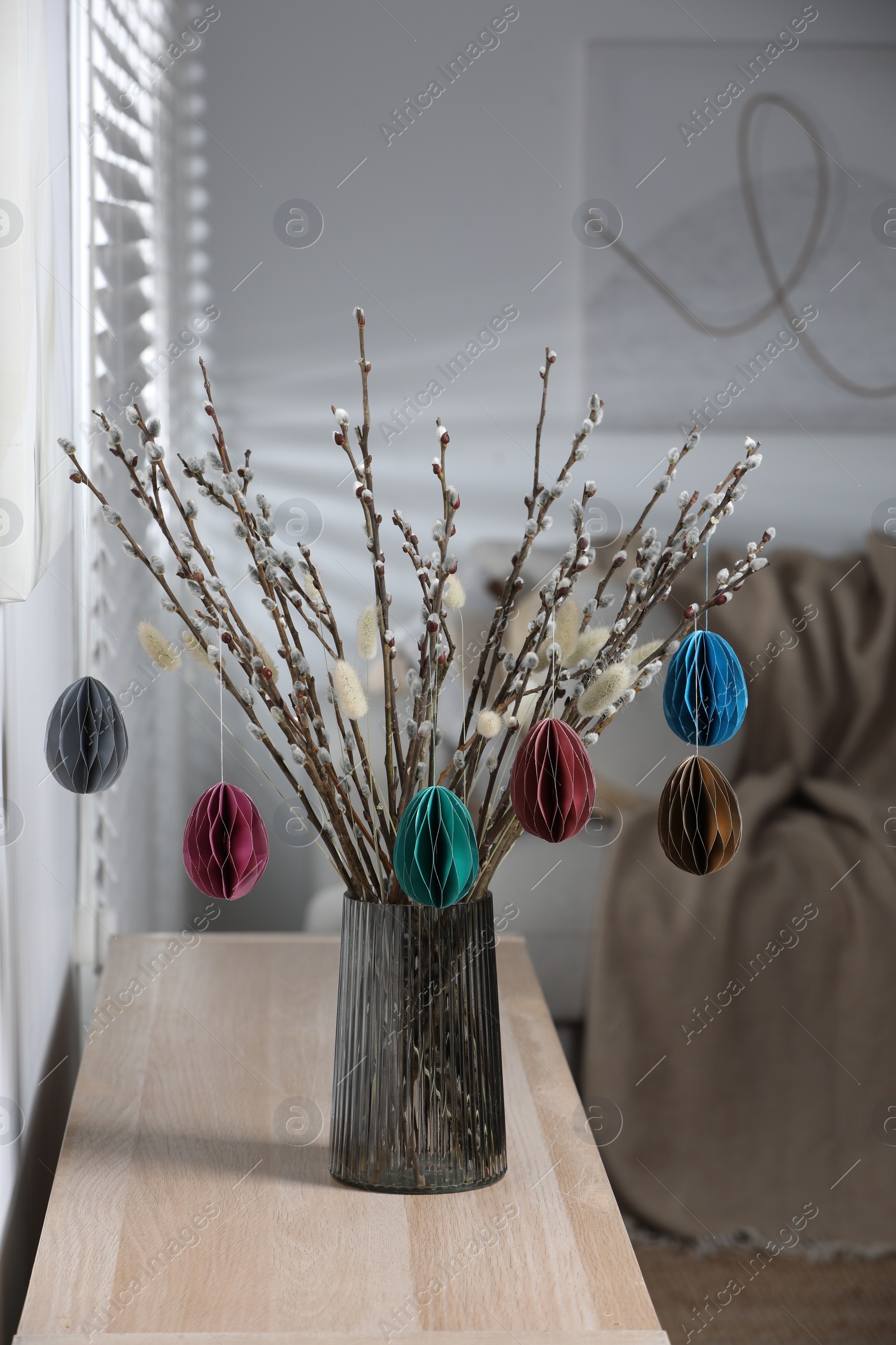 Photo of Beautiful pussy willow branches with paper eggs in vase on wooden table at home. Easter decor