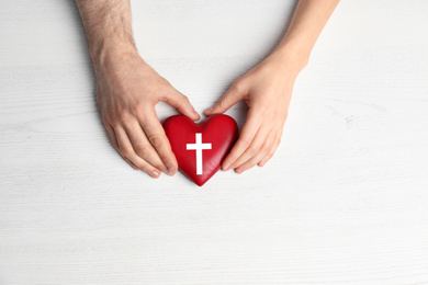 Couple holding heart with cross symbol on white wooden background, top view. Christian religion